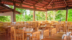 Un vaste espace extérieur aménagé pour les repas, avec des tables et des chaises en bois, entouré d’une végétation luxuriante et surmonté d’un toit recouvert de tuiles rouges.