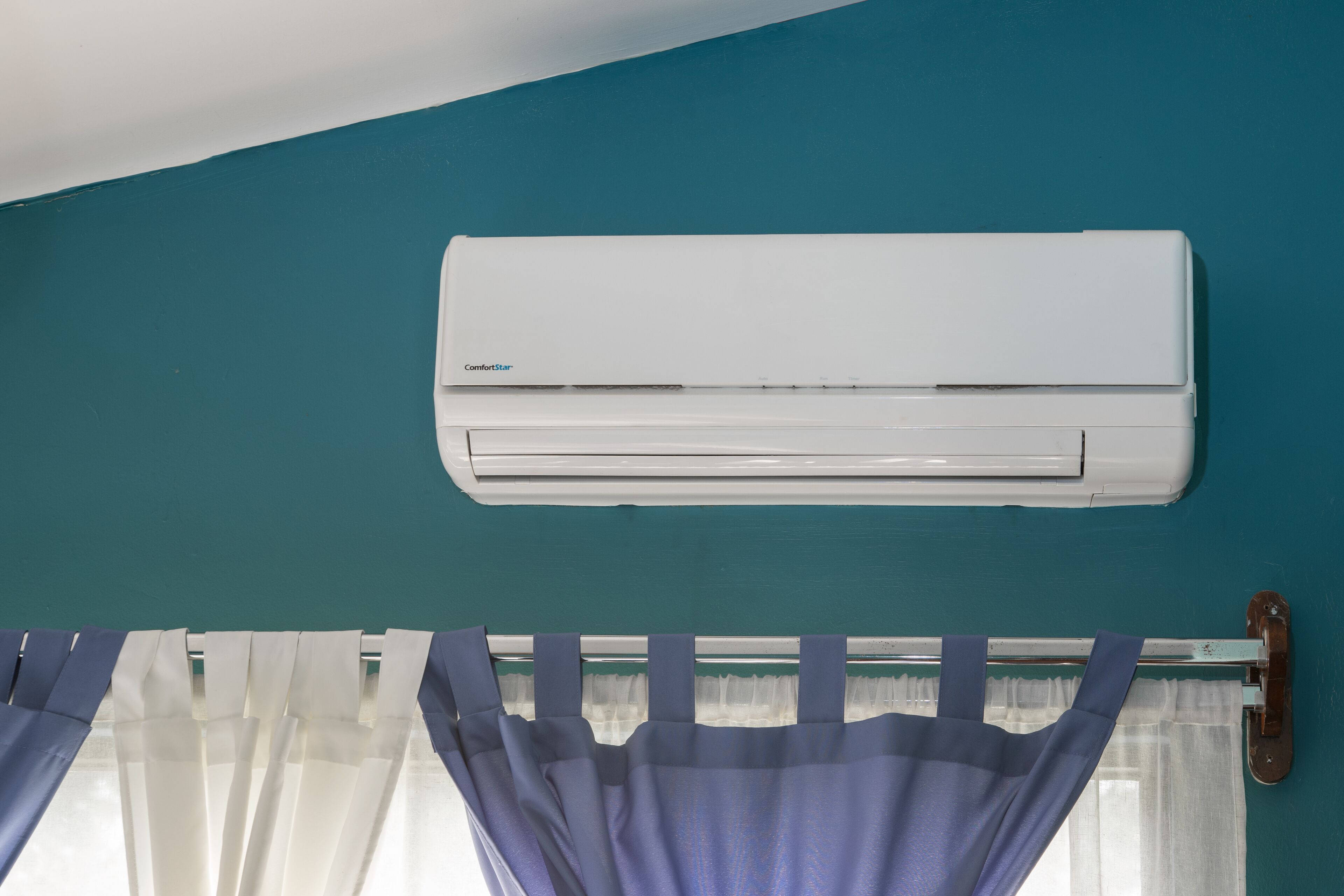A white air conditioner mounted on a teal wall above a window with purple curtains.