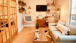 A cozy living room with a wooden coffee table, a sofa, and a TV mounted on the wall. There are potted plants on shelves and a hanging chair.