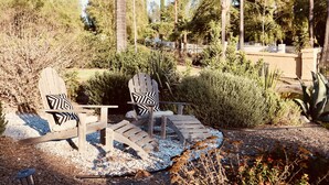 Two Adirondack chairs with patterned cushions, a small table, and a garden setting with various plants and a cactus.