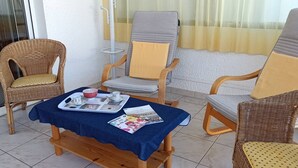 Une terrasse ensoleillée avec des chaises en osier, une table en bois recouverte d’une nappe bleue et une fenêtre ornée de rideaux légers.