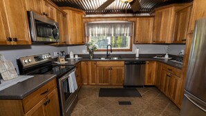 A kitchen with wooden cabinets, stainless steel appliances, a window with curtains, and a black countertop. - Casselman Cabin Retreat (Confluence)