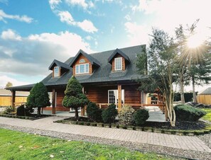 Une maison en bois à deux étages, dotée d’un toit en pente, d’un porche couvert et d’un jardin bien entretenu avec des arbustes taillés et un petit arbre.