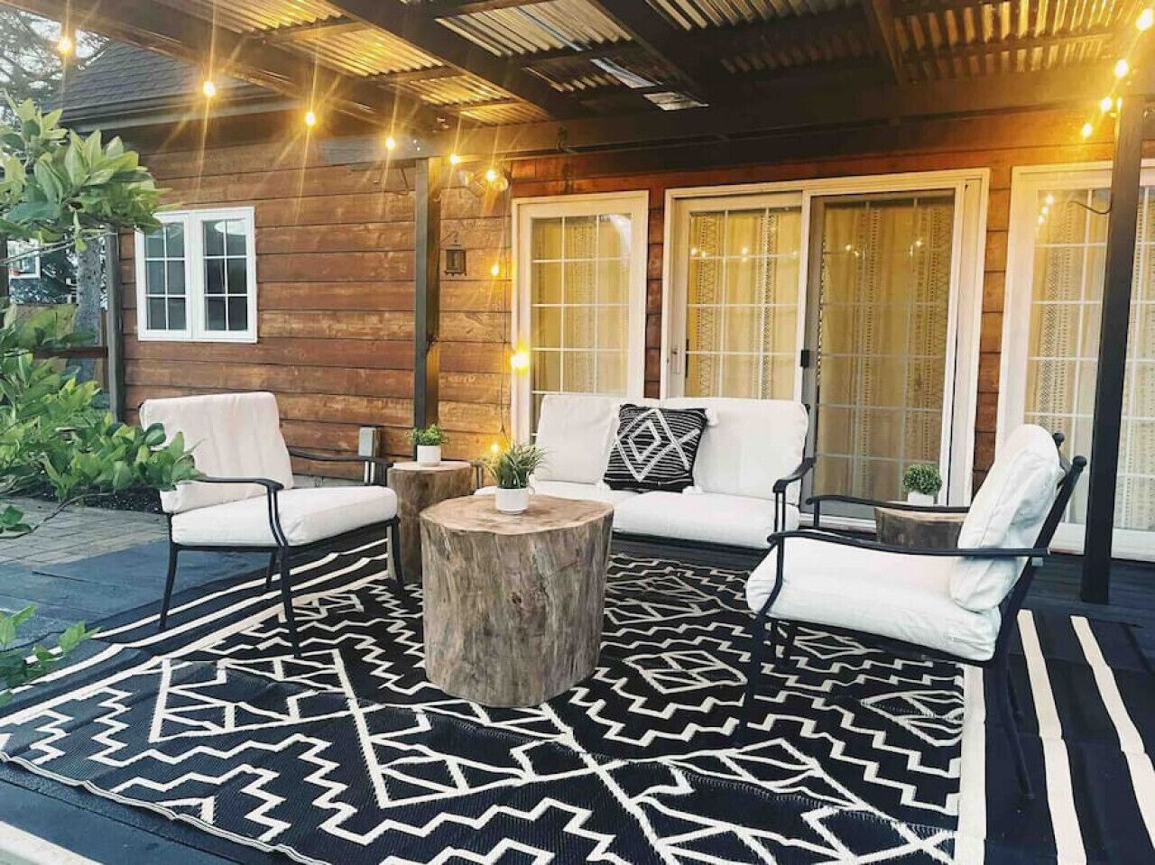 An outdoor seating area with a round wooden table, two white cushioned chairs, and a patterned rug.