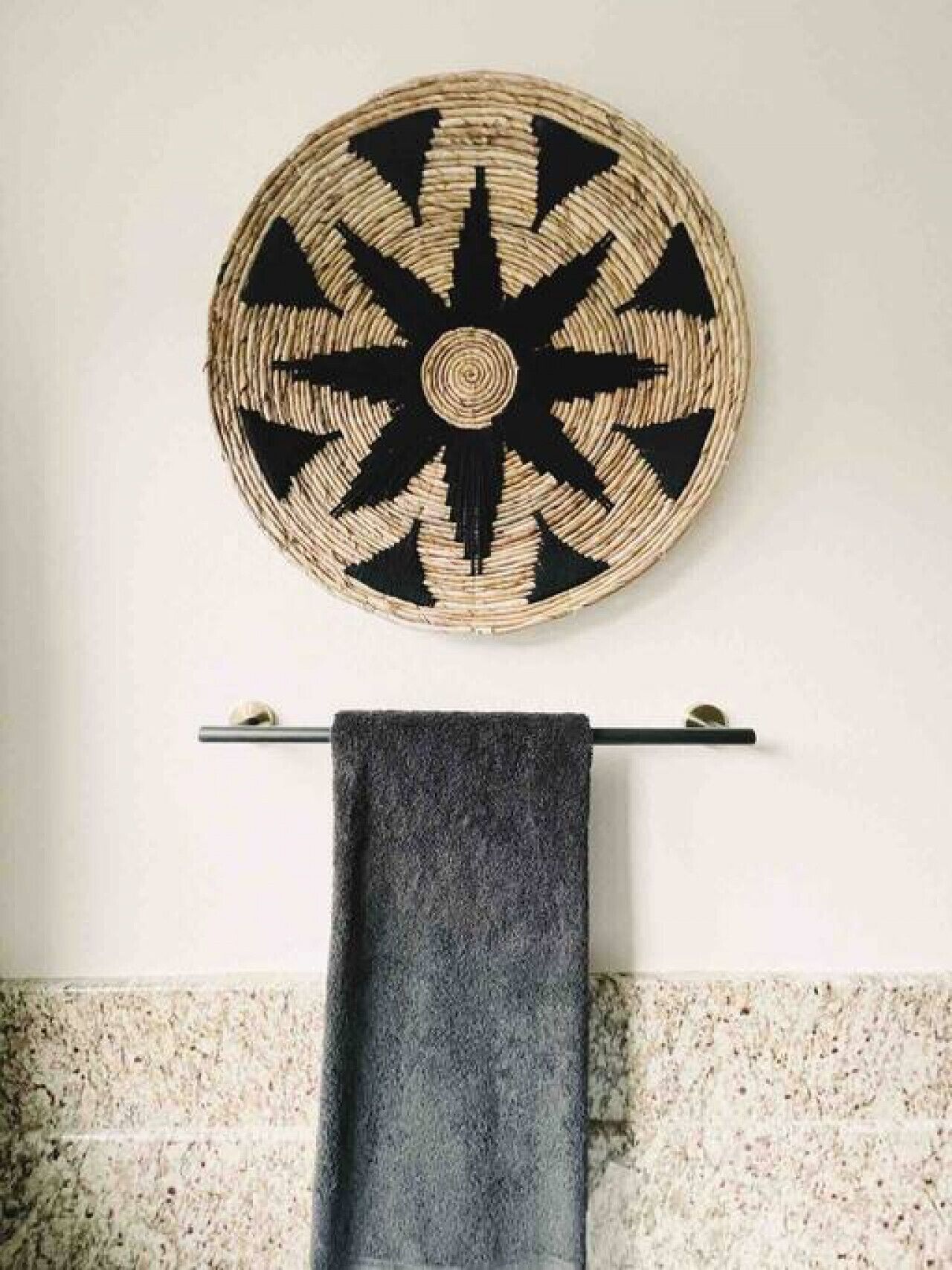 A circular woven wall hanging with a black and beige pattern, a towel hanging on a metal rod, and a speckled tile wall.