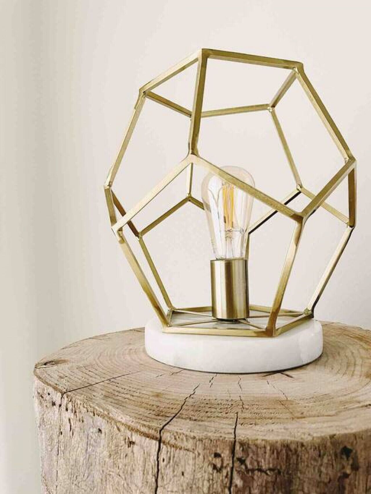 A geometric metal light fixture with a clear glass bulb on a white base, placed on a wooden stump.