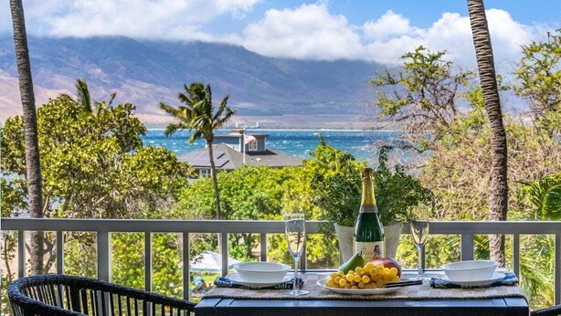 Une table avec une bouteille de champagne, un verre et une assiette de fruits, placée sur un balcon donnant sur une vue côtière, avec des palmiers et des montagnes en arrière-plan.