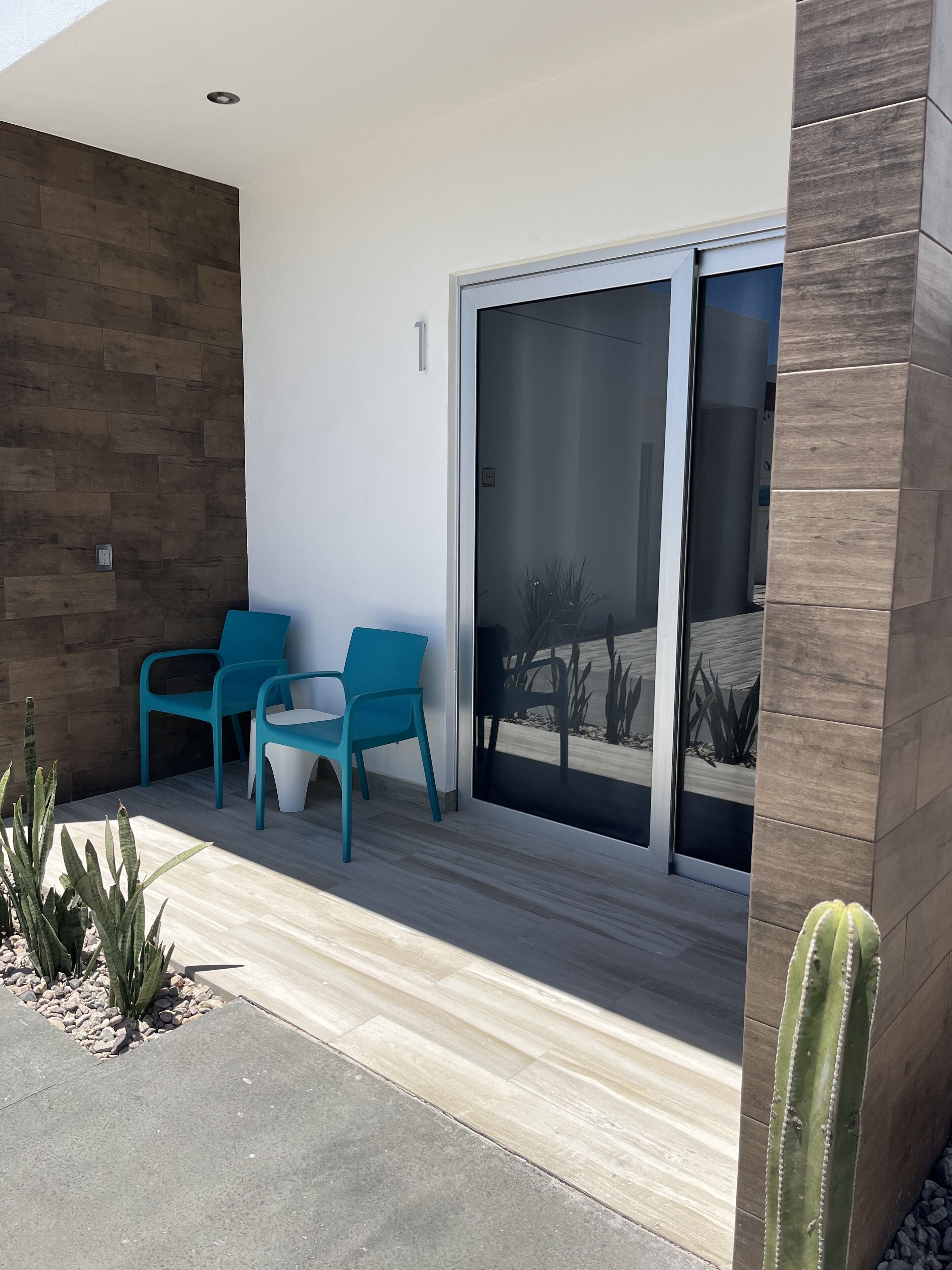 Uma área de pátio moderna com duas cadeiras turquesa e uma mesa, um painel de madeira e uma porta de vidro que dá para um jardim.