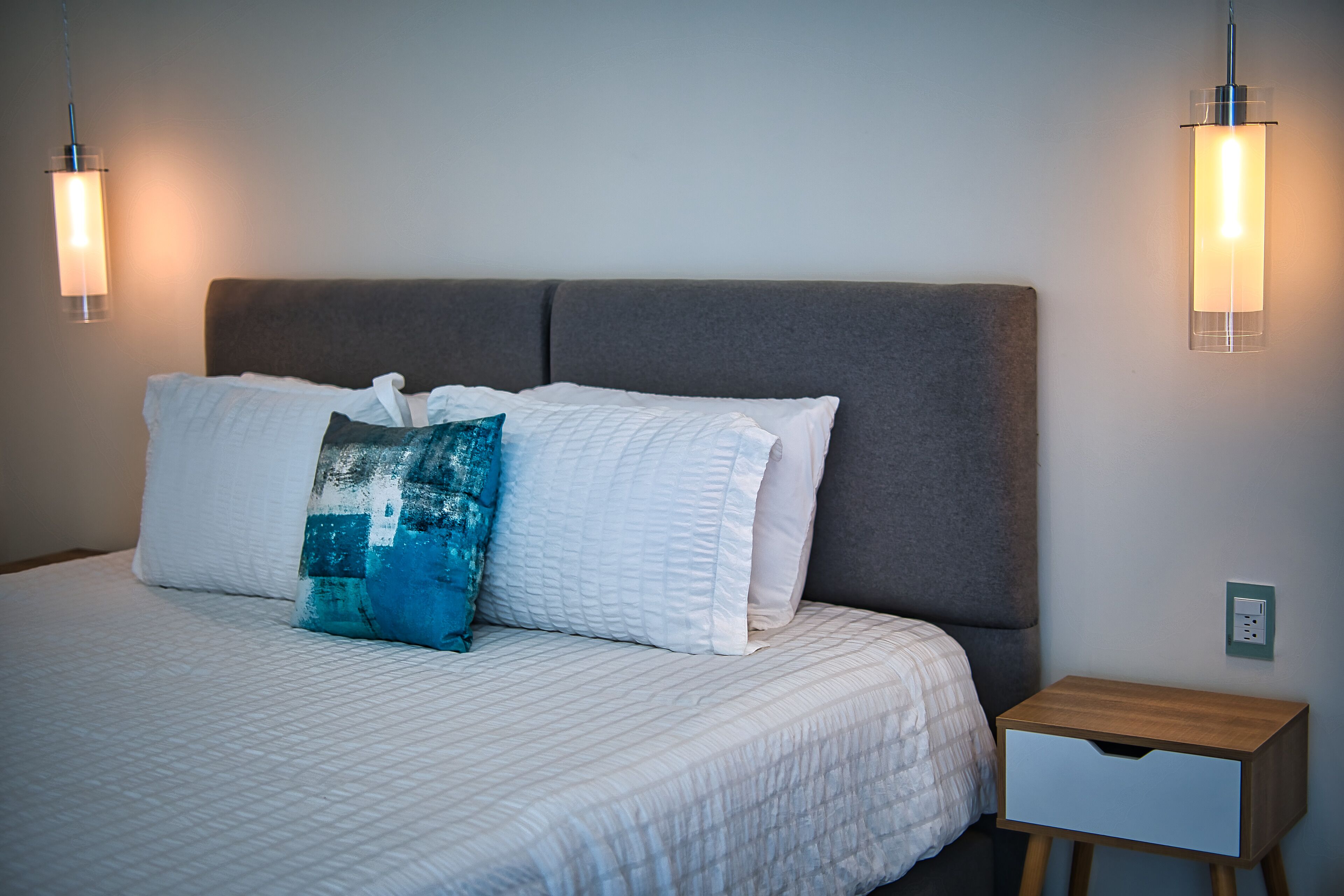 A neatly made bed with white bedding and a blue pillow, a wooden bedside table with a drawer, and two wall-mounted light fixtures.