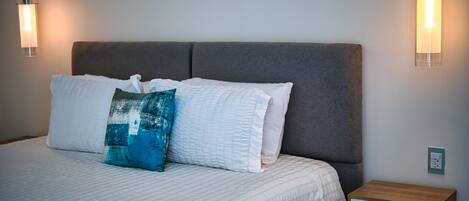Una cama bien hecha con ropa blanca y una almohada azul, una mesita de noche de madera con un cajón, y dos lámparas fijadas a la pared.