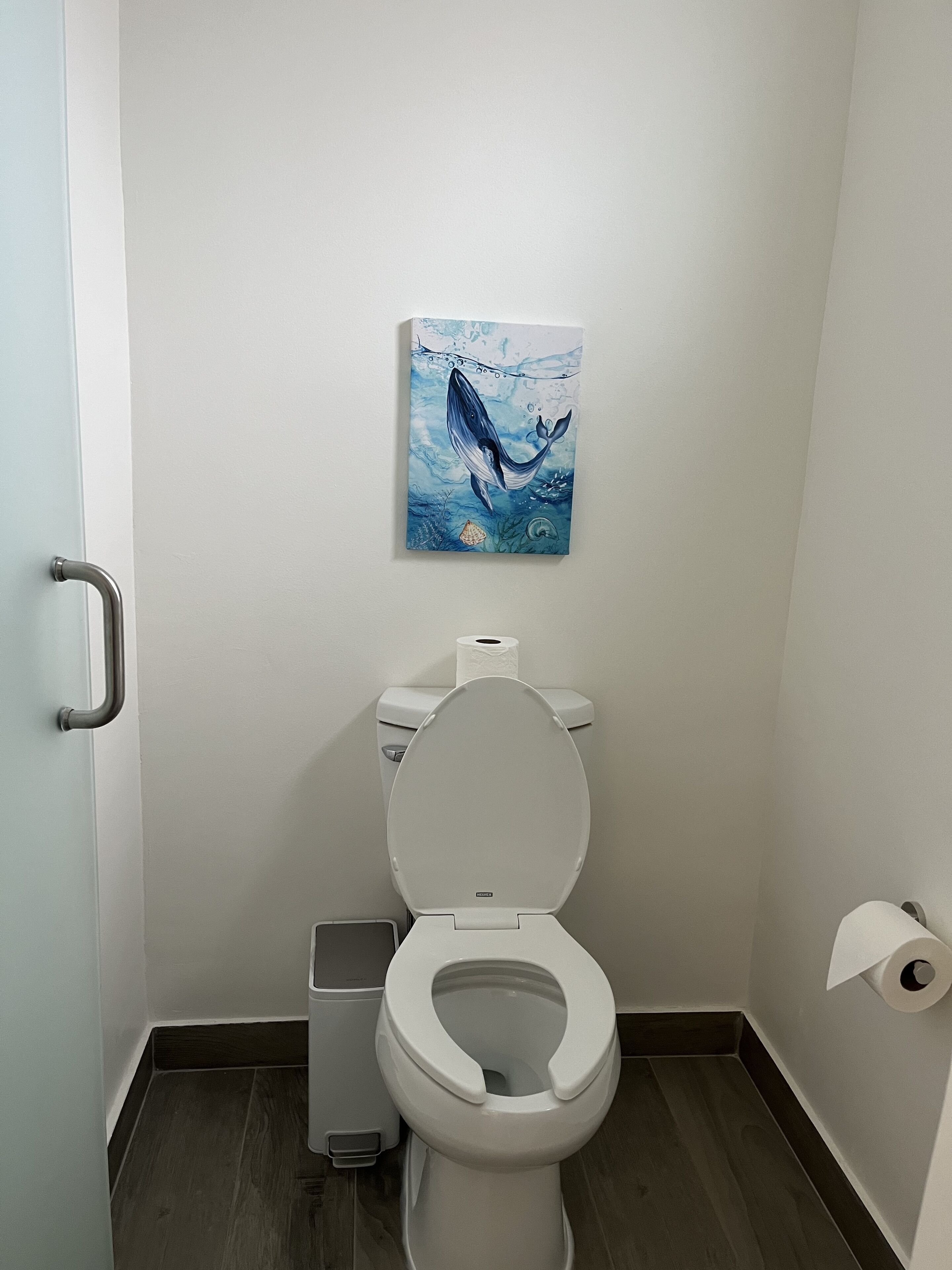 Une salle de bain équipée de toilettes, d’une barre d’appui, d’un porte-rouleau de papier toilette fixé au mur et d’un tableau représentant une baleine.