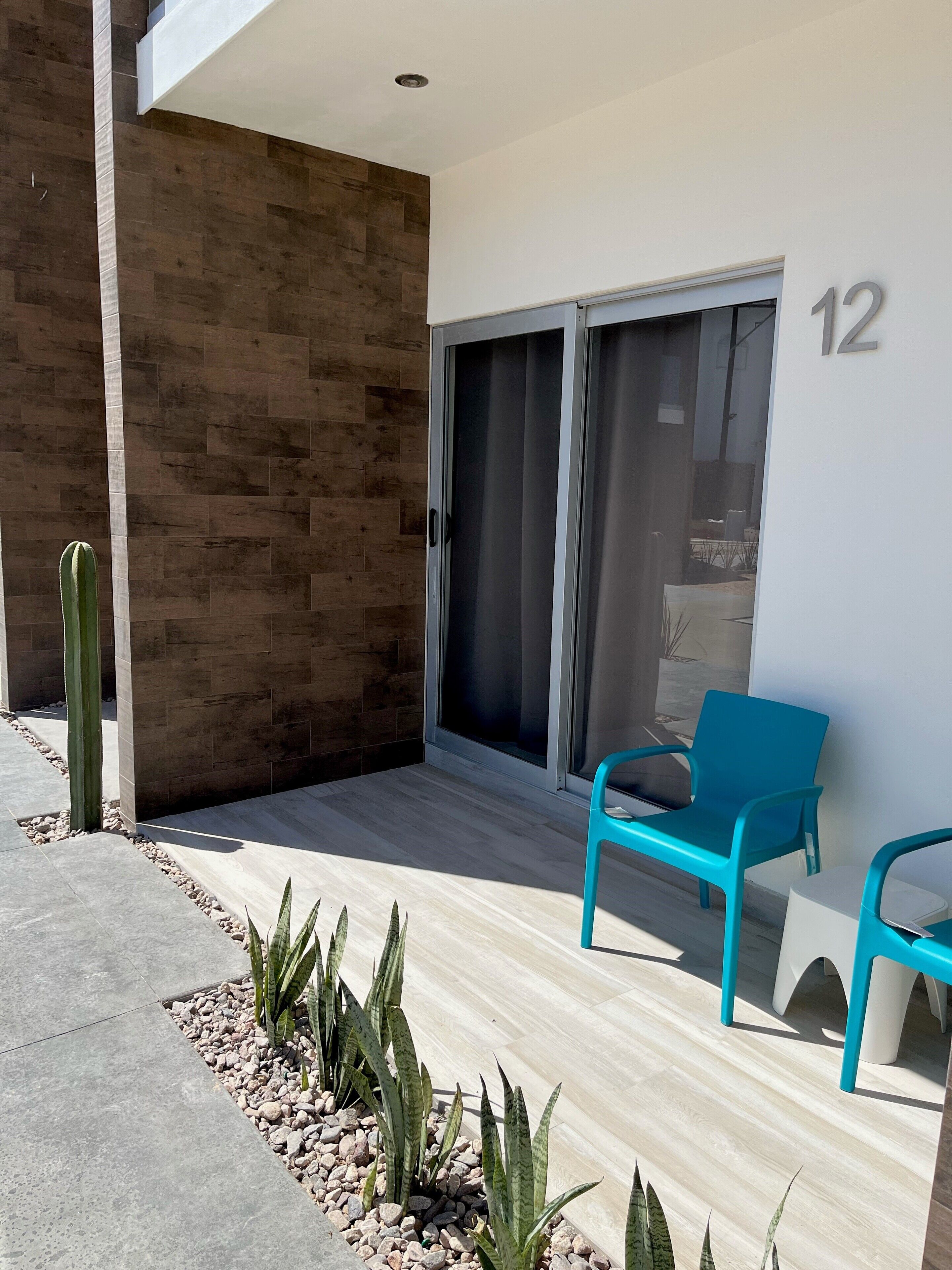Un'area esterna moderna con una porta a vetri scorrevoli, un cactus e due poltrone color turchese.