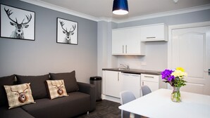 A modern living room with a dark brown sofa, two framed deer pictures, a white dining table with a vase of flowers, and a kitchen area with white cabinets and a sink. - St Andrews Guest House (Irvine)