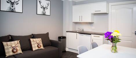 A modern living room with a dark brown sofa, two framed deer pictures, a white dining table with a vase of flowers, and a kitchen area with white cabinets and a sink.