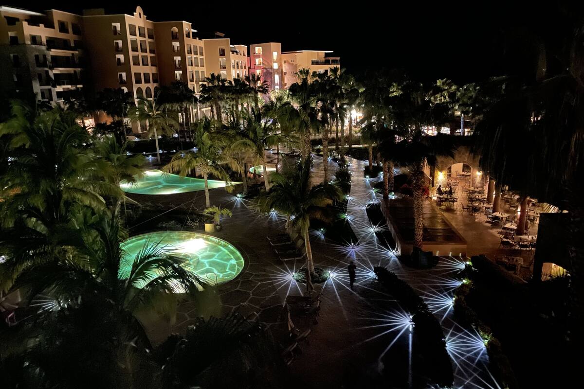 Vista nocturna de un resort con varias edificaciones, palmeras y senderos iluminados.