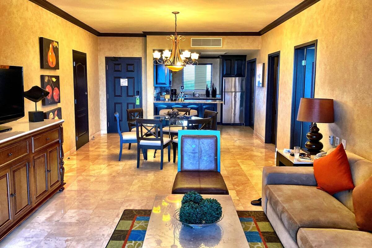 Amplia sala de estar con televisor, sofá y mesa de centro. Se ve una cocina al fondo, junto con un comedor.