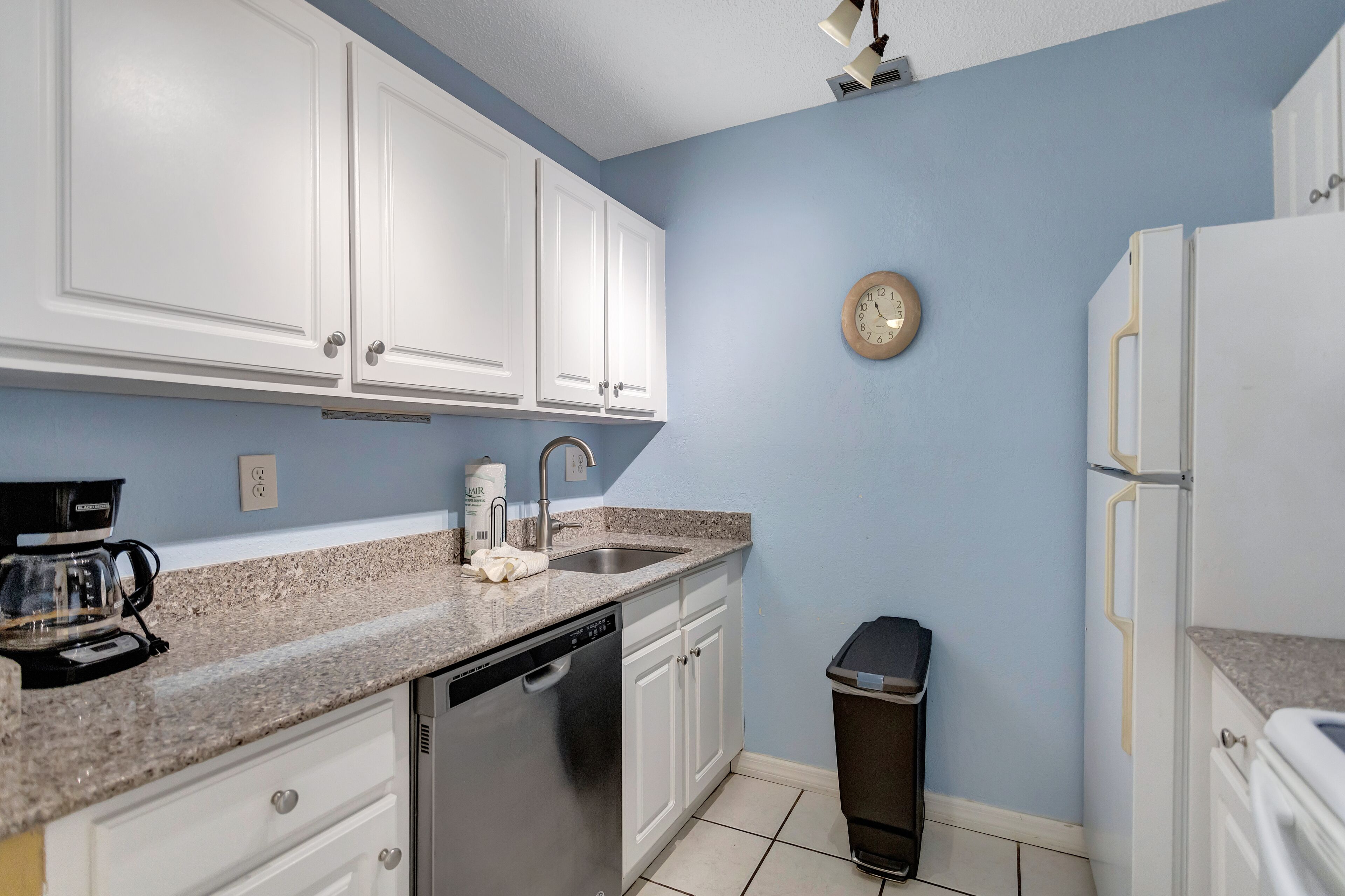 A kitchen with white cabinets, a granite countertop, a coffee maker, a dishwasher, a sink, a trash can, and a clock on the wall.