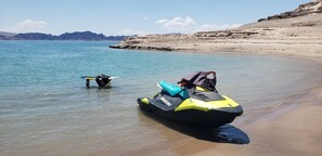 Ein Jetski und ein Surfbrett an einem sandigen Strand mit ruhigem Wasser und Bergen im Hintergrund.