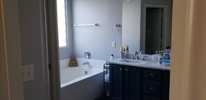 Ein Badezimmer mit einer Badewanne, zwei Waschbecken und einem großen Spiegel. Auf dem Waschtisch liegen Pflegeprodukte und über der Badewanne befindet sich ein Handtuchhalter.