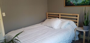 Ein Einzelbett mit Holzkopfstütze, ein kleiner runder Tisch mit einem Blumentopf daneben und ein an der Wand hängendes Bild.