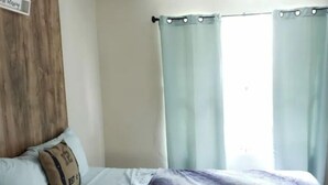 Un dormitorio con cama, almohadas, una colcha y cortinas claras.
