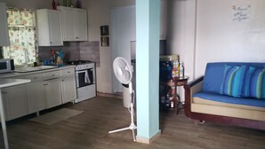 Cocina compacta con armarios blancos, estufa y microondas. Hay un ventilador en la esquina y un sofá azul con cojines a rayas en la sala.