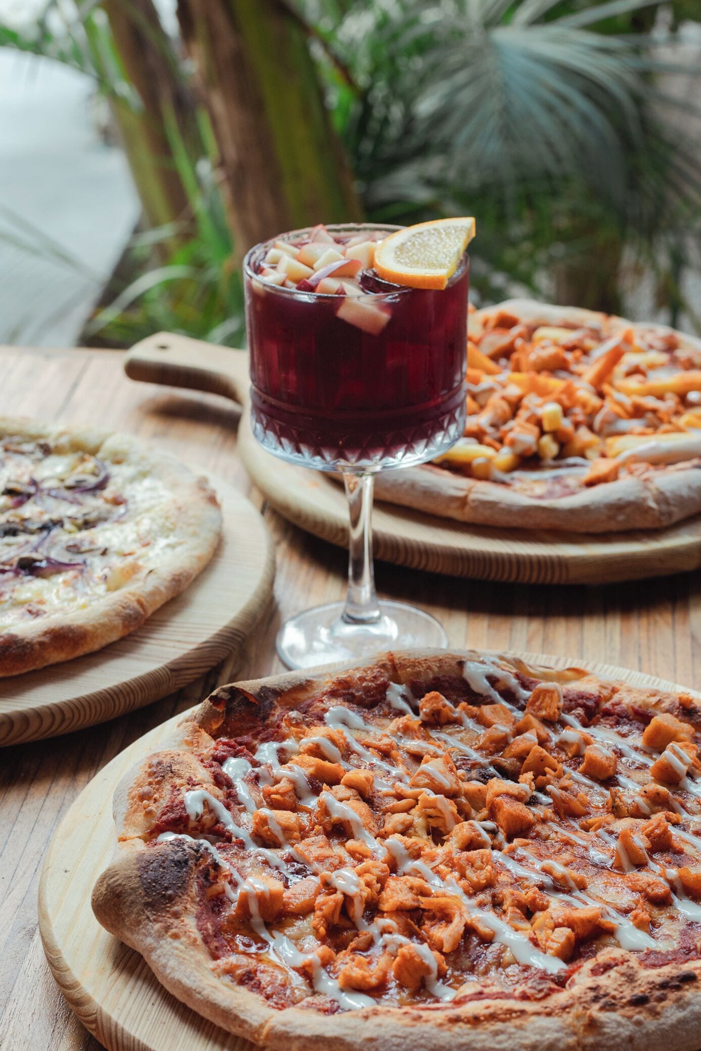 Three pizzas on wooden boards with a glass of red beverage and a slice of orange on the rim.