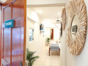 A hotel room with a wooden door, a bed with a white and gray bedspread, a small table with a white tablecloth, a potted plant, and a wall-mounted mirror with a decorative frame. - Zulu Surf Hotel (Tamarindo)