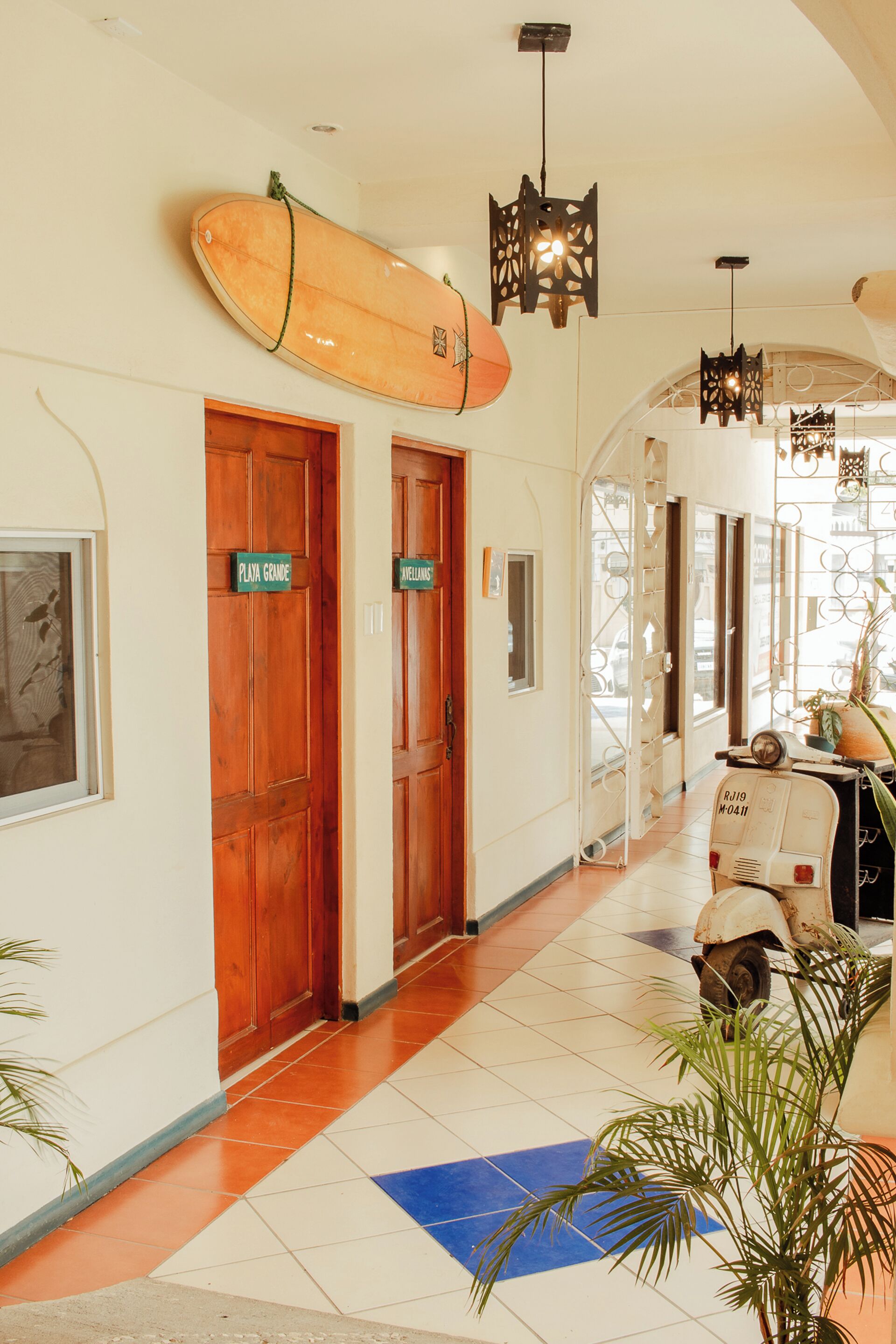 Een hotelgang met houten deuren met de aanduiding 'Playa Grande' en 'Javellanes', een surfplank aan de muur en een vintage Vespa scooter.