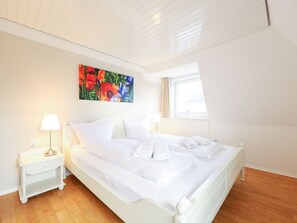 Quarto com uma cama grande, roupa de cama branca, mesa de cabeceira com abajur e um quadro floral colorido na parede.