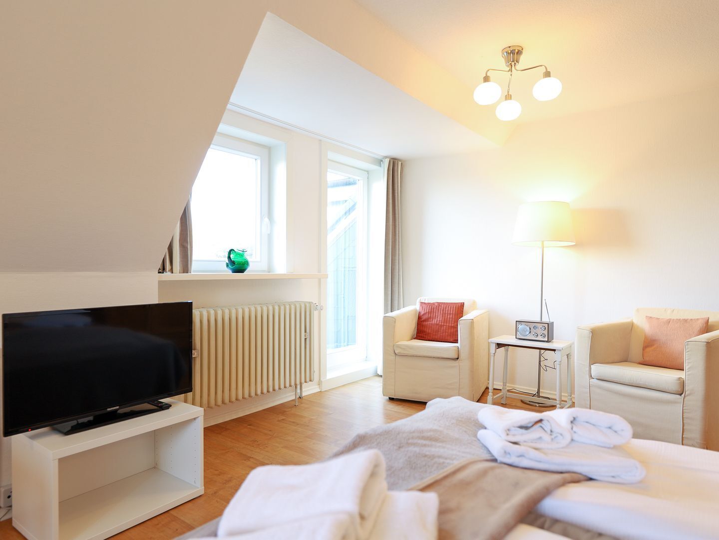 Una habitación de hotel moderna con televisión de pantalla plana, un radiador, una ventana con cortinas, una lámpara y un sofá.