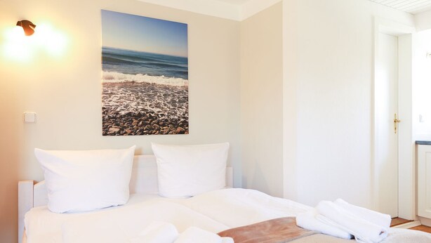 Una habitación de hotel con una cama, almohadas blancas, una colcha color beige y un cuadro de una playa en la pared.