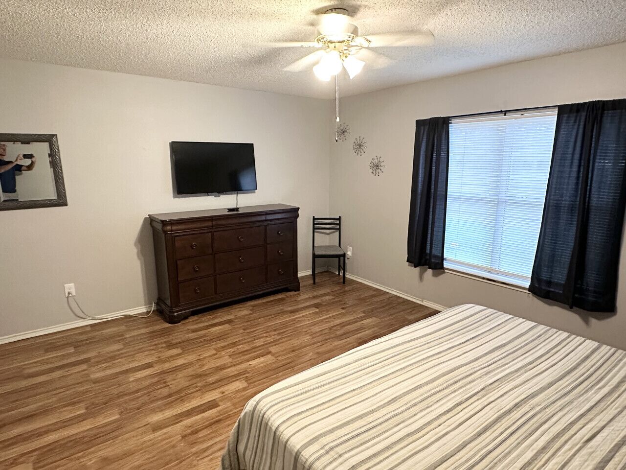 Une chambre à coucher comprenant un lit, une commode, un téléviseur, un ventilateur de plafond et une fenêtre avec des rideaux.