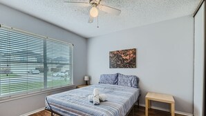Un dormitorio con una cama, un ventilador de techo, una ventana con persianas, una mesita de noche y un pequeño banco.