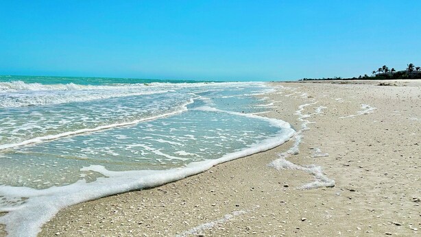 A beach with clear water, a sandy shore, and a distant building. - Gulfside Place #107 ~ 3br/3ba - Gulf front living at its finest (Sanibel)