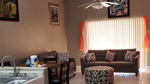Ein Wohnzimmer mit Sofa, Hocker, Esstisch und Stühlen. Es gibt einen Deckenventilator, ein Bild an der Wand und ein Fenster mit Jalousien.