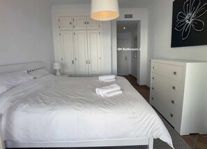 Une chambre à coucher avec un grand lit, du linge de lit blanc, une table de chevet avec une lampe, une commode et une porte donnant sur une salle de bain.