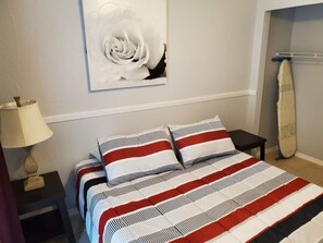 Een bed met gestreept beddengoed, een aan de muur hangend kunstwerk, een lamp en een droogrek.