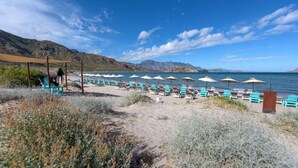 En strand med rader av blå stolar och vita parasoller, ett sandigt område med sparsam växtlighet och en fri utsikt över havet och de avlägsna bergen.