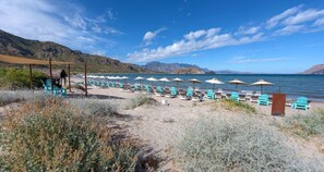 En strand med rader av blå stolar och vita parasoller, ett sandigt område med sparsam växtlighet och en fri utsikt över havet och de avlägsna bergen.