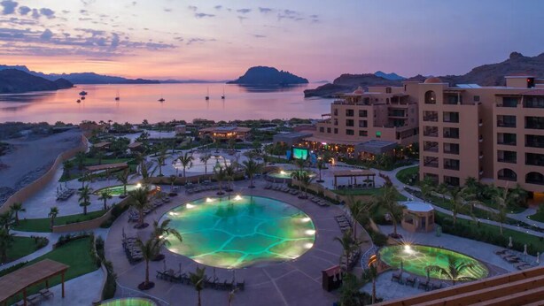 Ett semesterhotell med en stor pool, flera byggnader och utsikt över havet i skymningen.