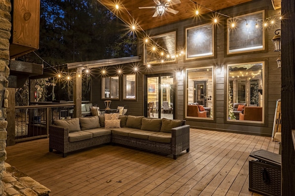A wooden deck with a wicker sofa and a table, string lights, and large windows.