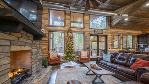 A cozy cabin interior with a stone fireplace, leather seating, a Christmas tree, and large windows offering views of the forest. - Cabin Therapy - Luxury Cabin Near ATV Trails (Broken Bow)