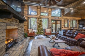 A cozy cabin interior with a stone fireplace, leather seating, a Christmas tree, and large windows offering views of the forest.