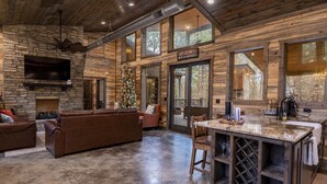 A rustic cabin interior with a stone fireplace, leather sofas, a wooden bar, and large windows offering views of the surrounding forest. - Cabin Therapy - Luxury Cabin Near ATV Trails (Broken Bow)