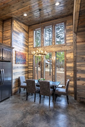 A rustic wooden interior with a dining area featuring a table and chairs, a refrigerator, and a large window with a view of the outdoors.