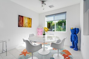 Un espace repas moderne avec une table ronde, des chaises blanches, un tapis coloré, une grande fenêtre et un ventilateur de plafond.