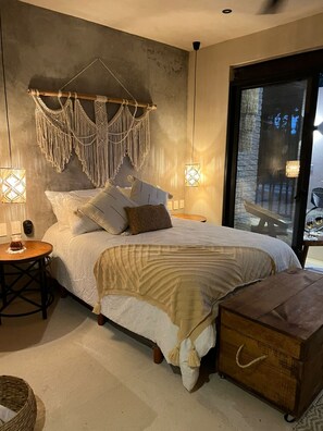 A bedroom with a bed, bedside tables, a macrame wall hanging, and a view of the outdoors through a glass door. - Luxury Studios (Tulum)