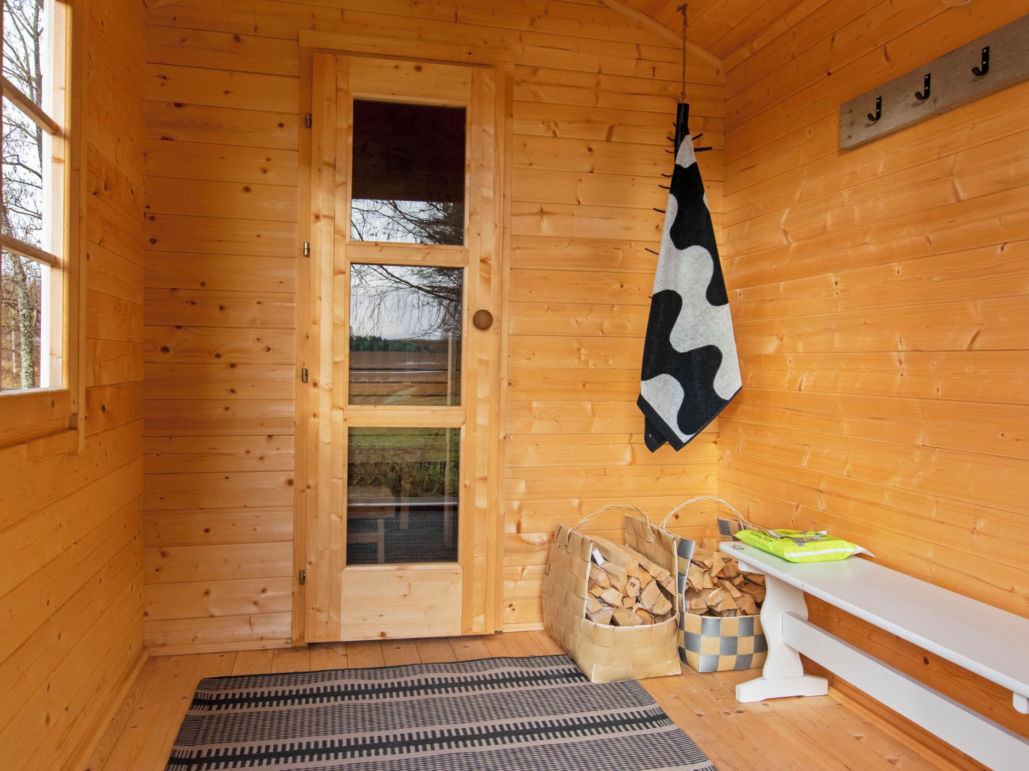 A wooden cabin interior with a door, a hanging cowhide, a bench, a woven basket, and a checkered rug.