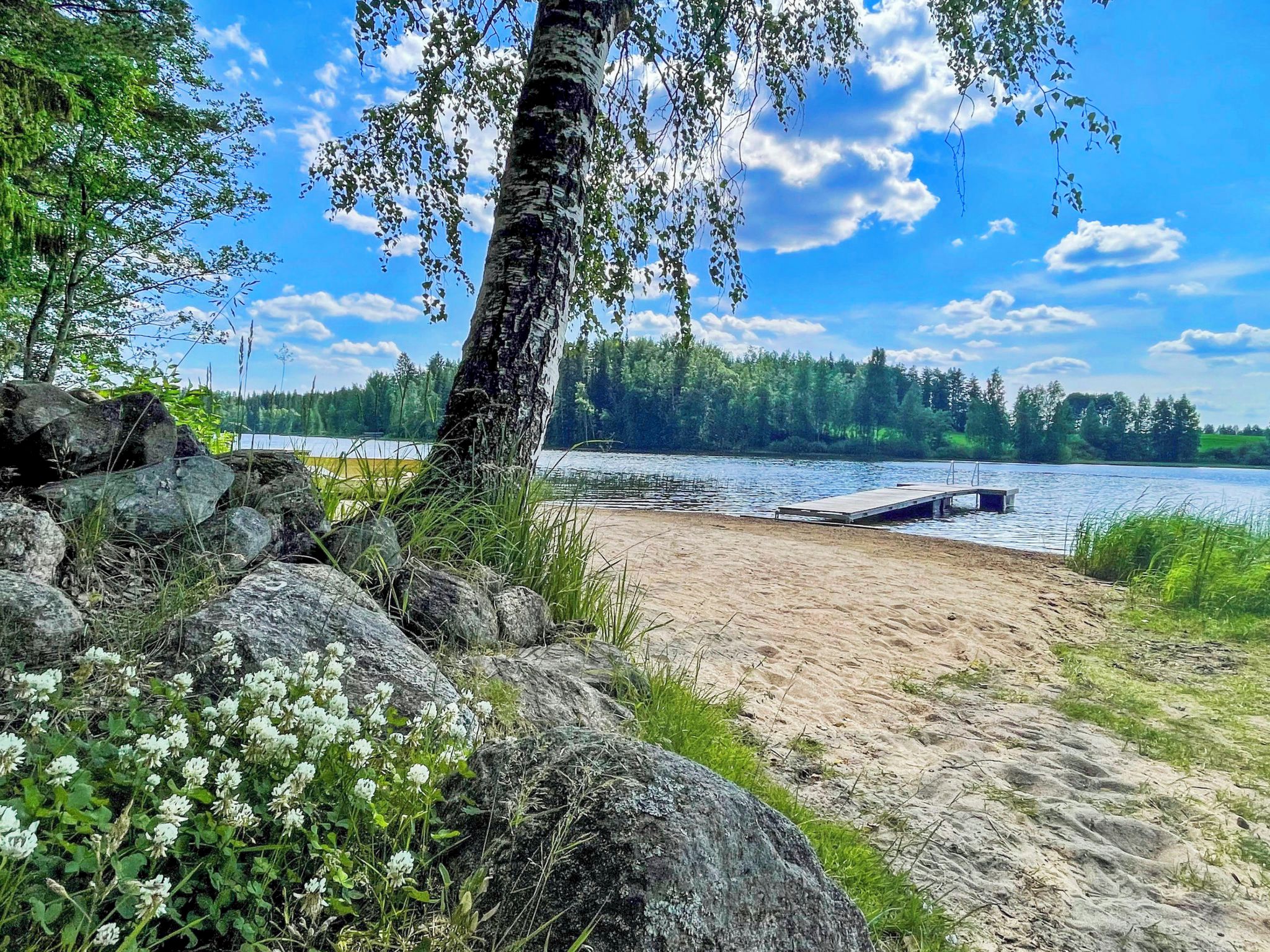 A sandy beach with a dock extending into a calm lake, surrounded by rocks and greenery.